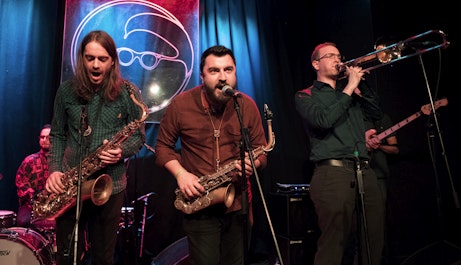 Three members of Funk Obligations performing live at Band on the Wall. Two are playing saxophones and singing into microphones, another is playing a trombone.