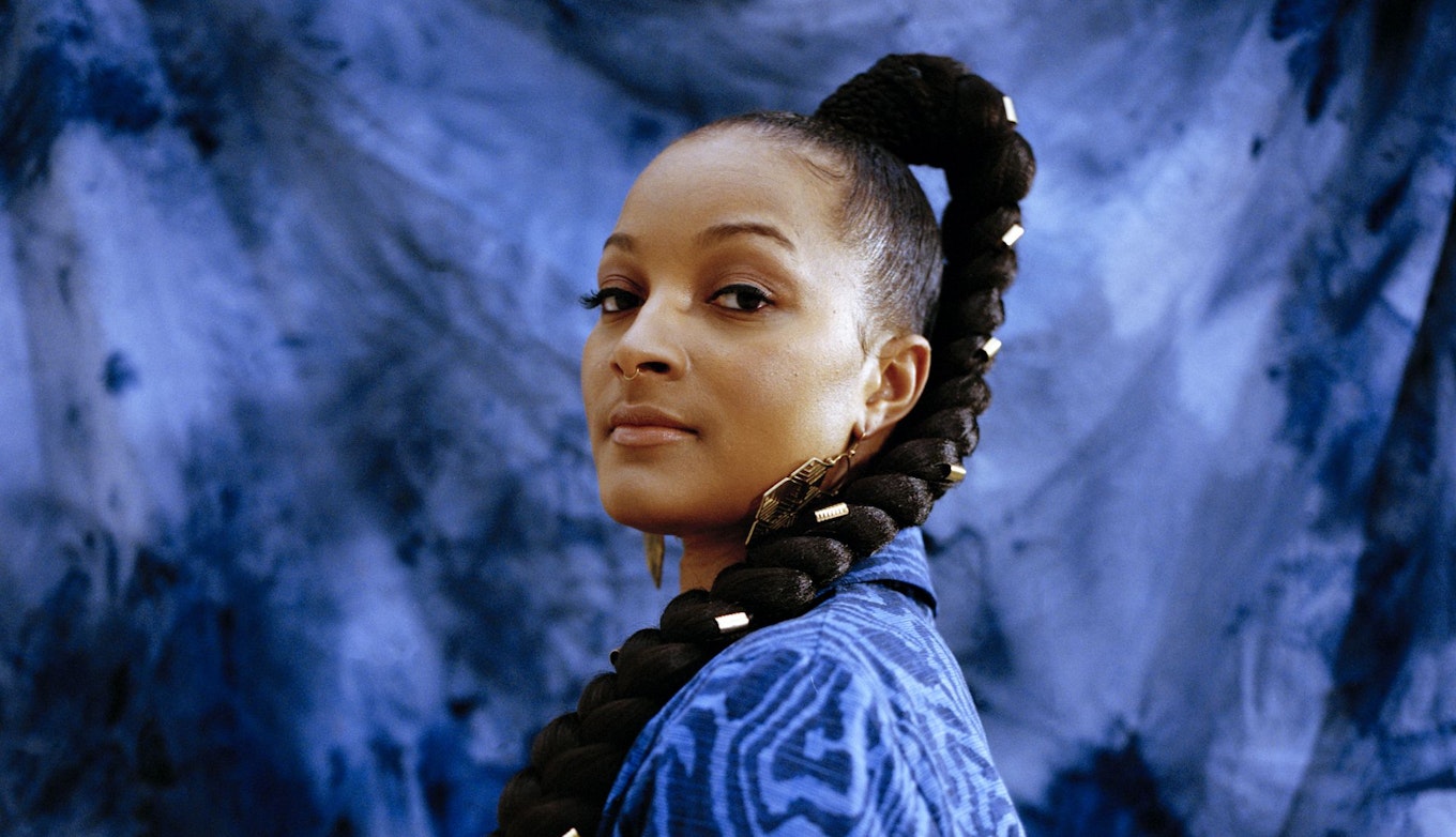 Nubya is wearing a wearing a blue patterned blazer and has gold details in their plait. Nubya looks directly at the camera and is standing in front of a blue tie dye background. 