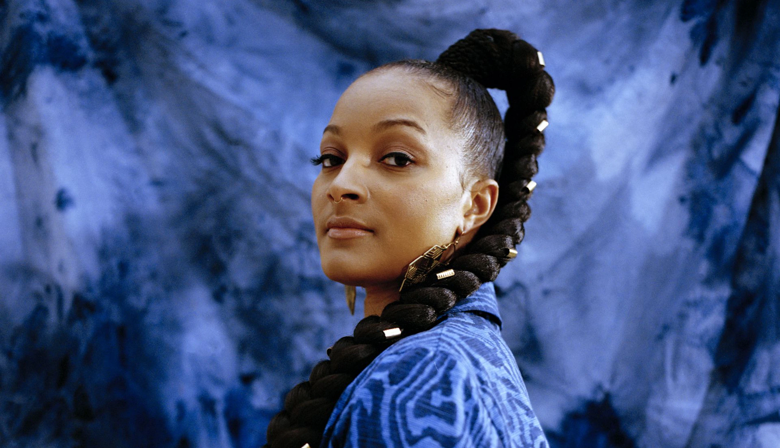 Nubya is wearing a wearing a blue patterned blazer and has gold details in their plait. Nubya looks directly at the camera and is standing in front of a blue tie dye background.