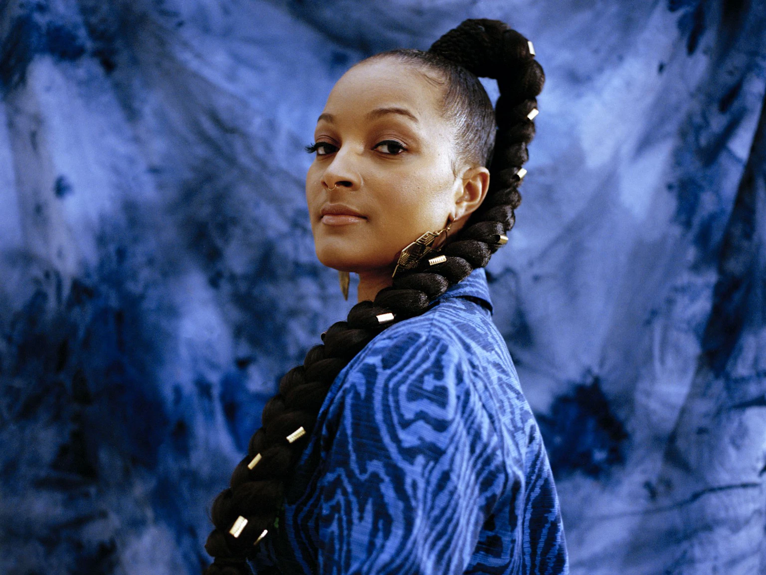 Nubya is wearing a wearing a blue patterned blazer and has gold details in their plait. Nubya looks directly at the camera and is standing in front of a blue tie dye background.