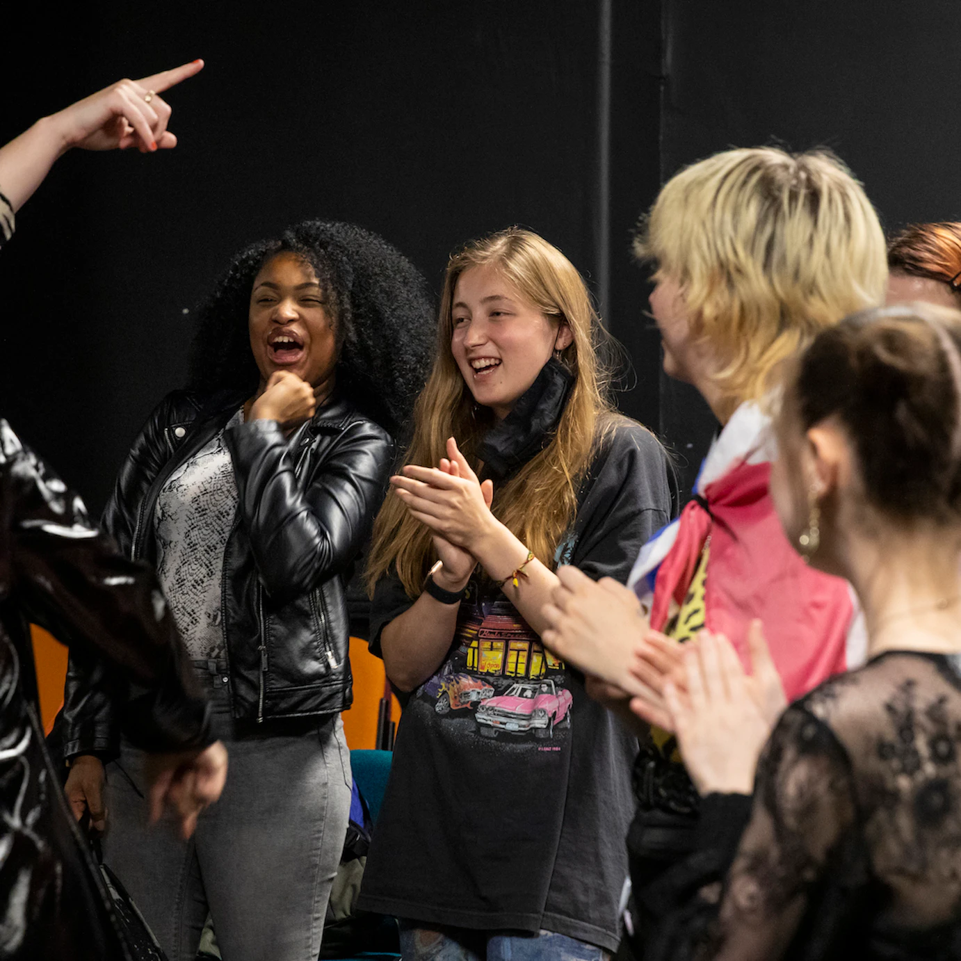 A group of eight young people are stood talking, laughing and clapping. They look happy and excited.