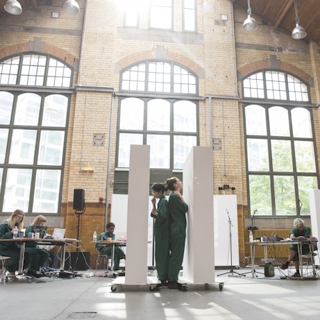 Residency artists in the Engine Hall in People's History Museum rehearsing. In the centre are two artists stood back to back between two white monoliths. Surrounding them artists sit at tables working on laptops.