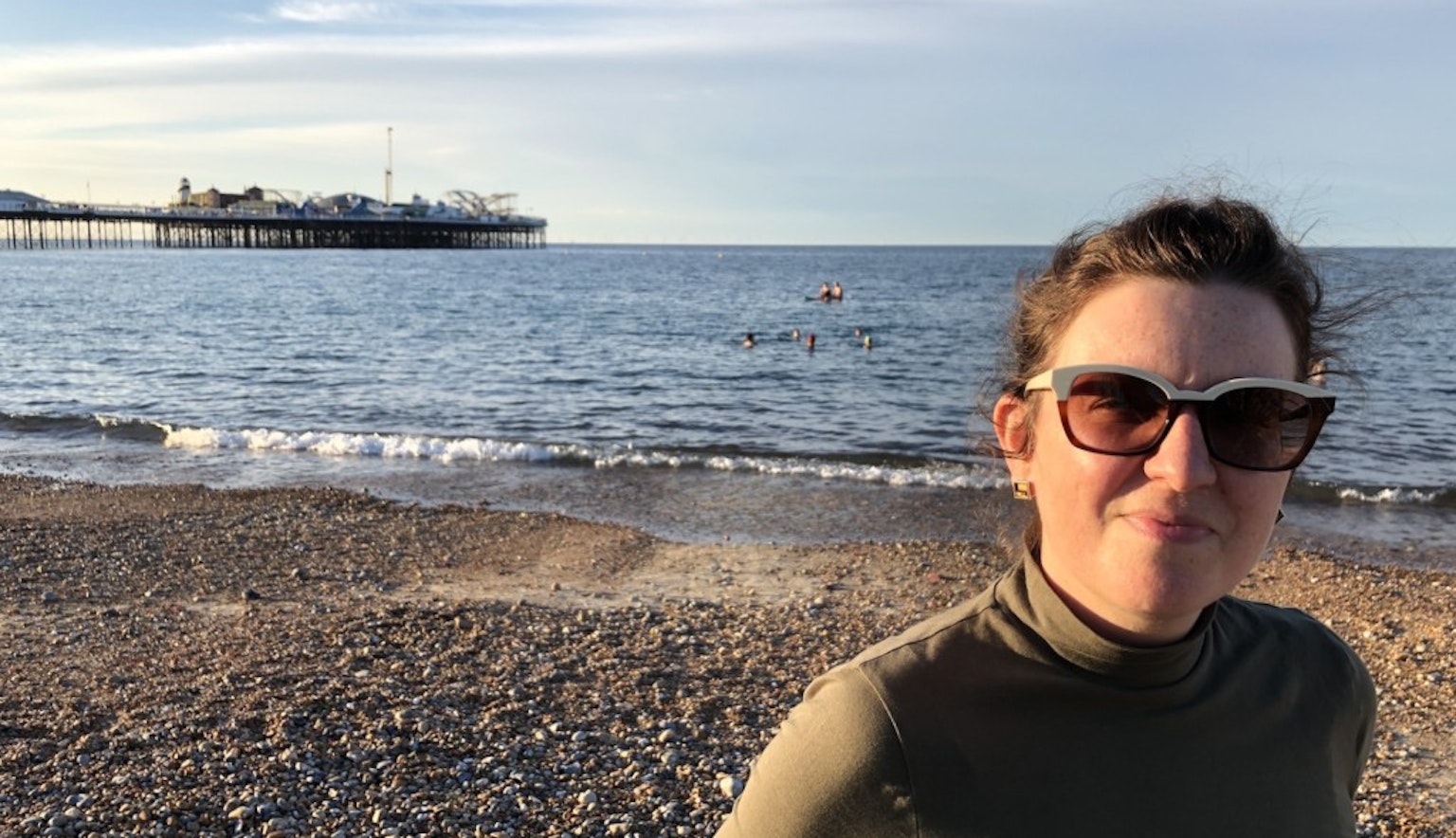 Anna Moulson stands to the right with sunglasses on looking at the camera. She is on a rocky beach. behind her is the ocean and a pier far off to the left.