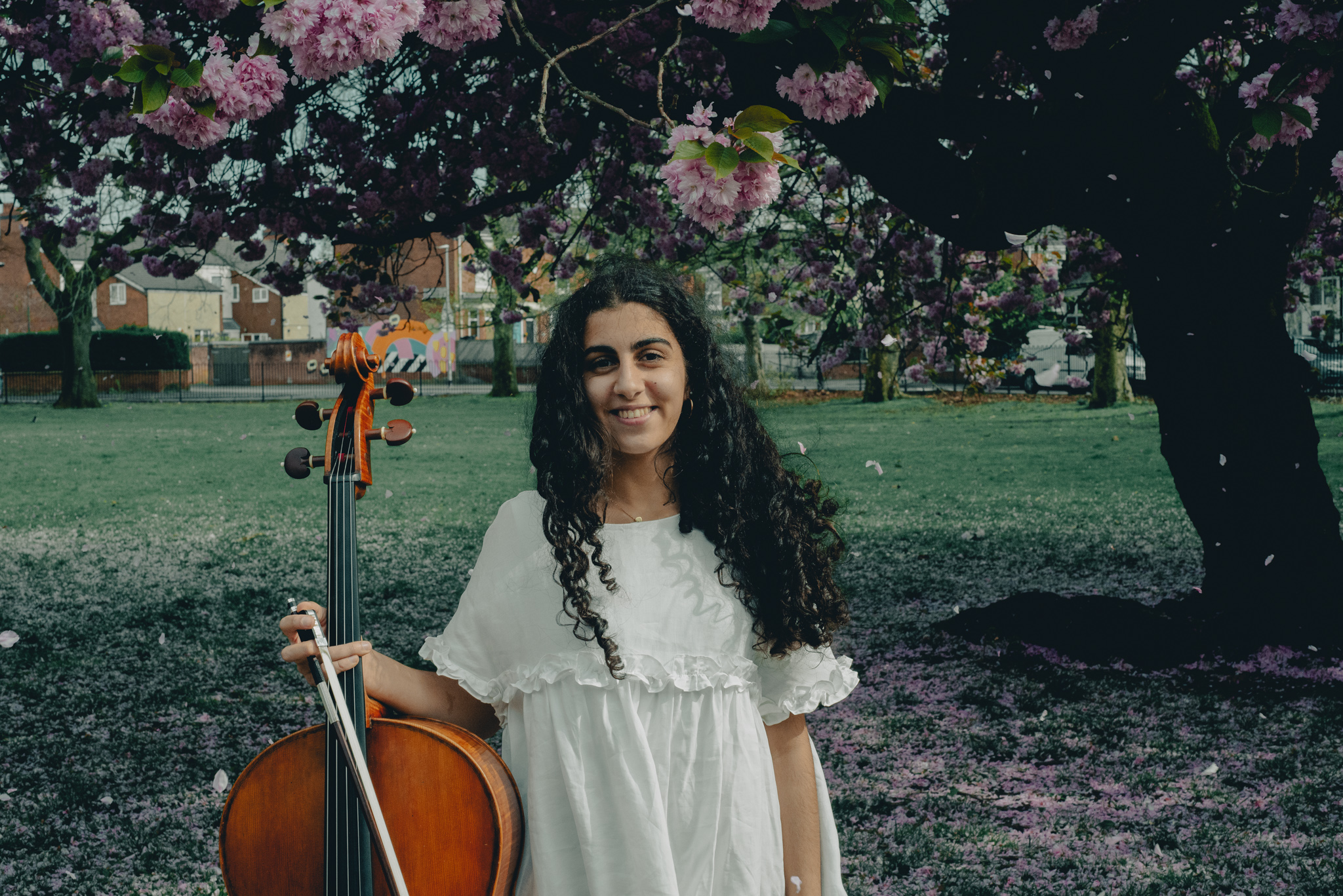 Hoda Jahanpour holding a cello, stood in front of a small park with blossom trees dotted around the outskirts.
