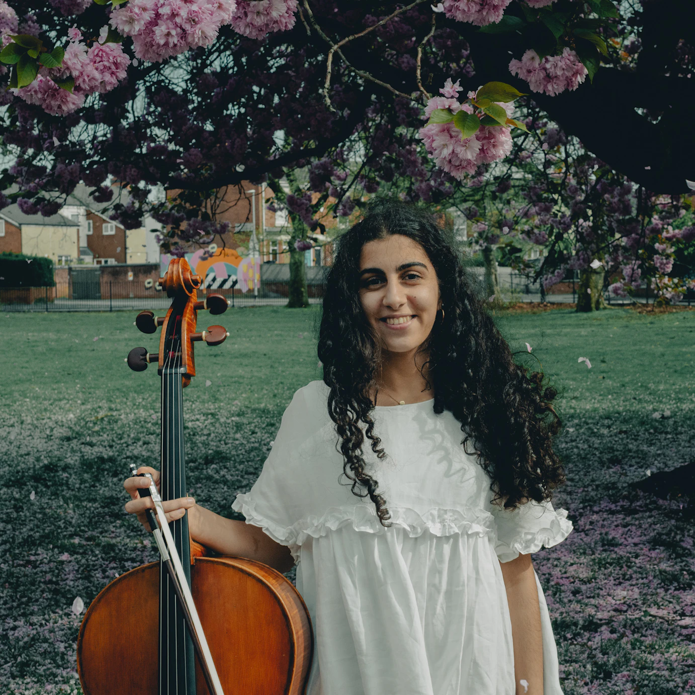 Hoda Jahanpour holding a cello, stood in front of a small park with blossom trees dotted around the outskirts.