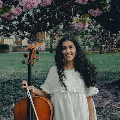 Hoda Jahanpour holding a cello, stood in front of a small park with blossom trees dotted around the outskirts.