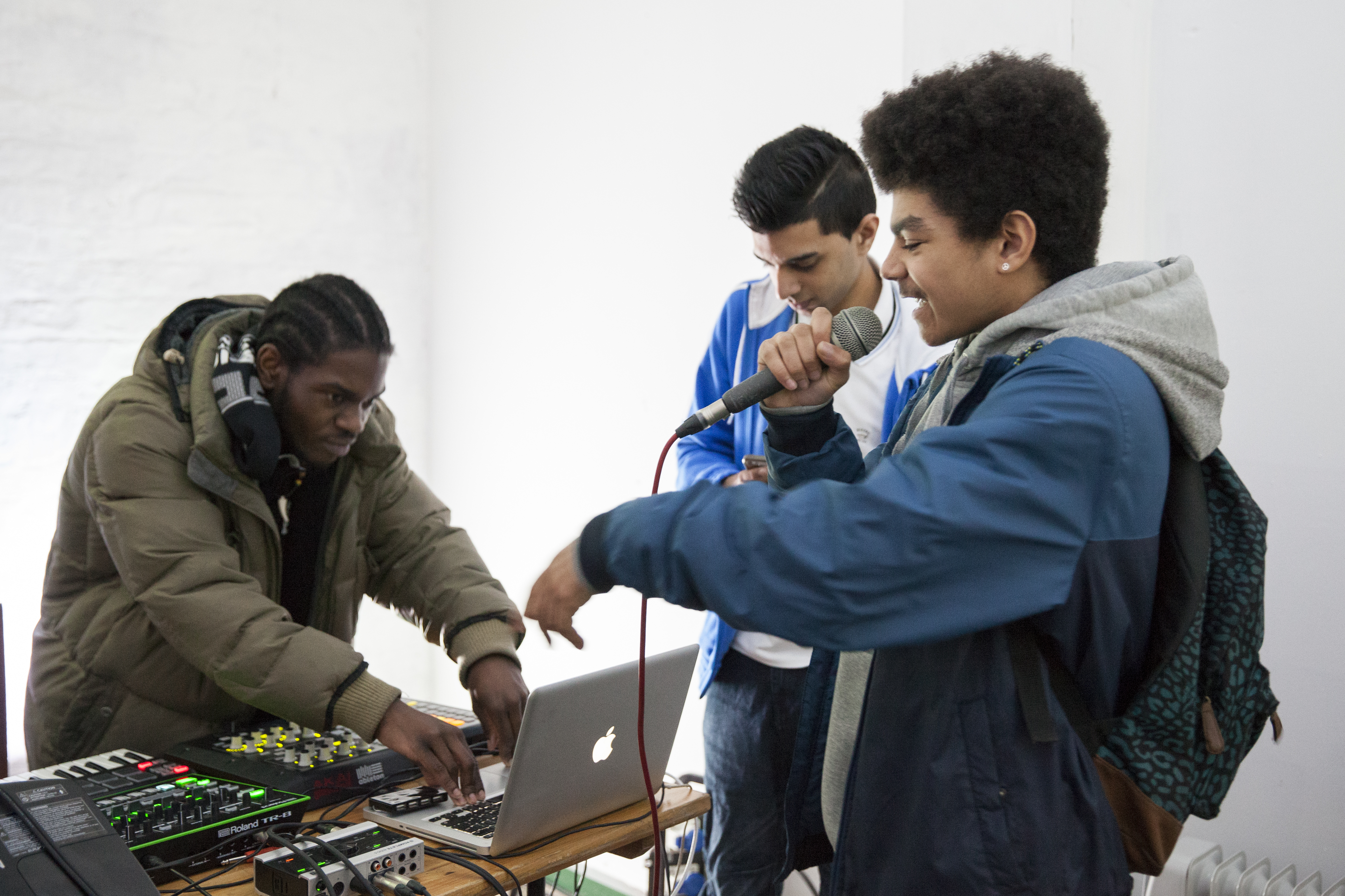 hree participants huddle around a production area. One is rapping into the microphone, another is mixing on the laptop and the other is looking at the laptop. 