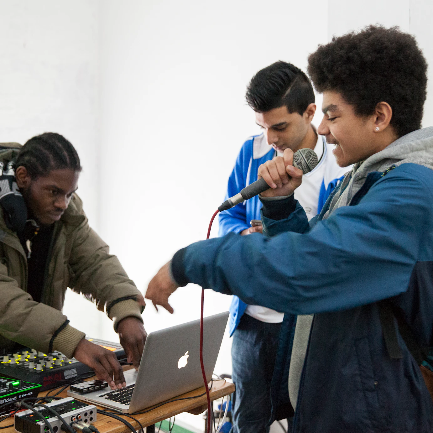 hree participants huddle around a production area. One is rapping into the microphone, another is mixing on the laptop and the other is looking at the laptop.