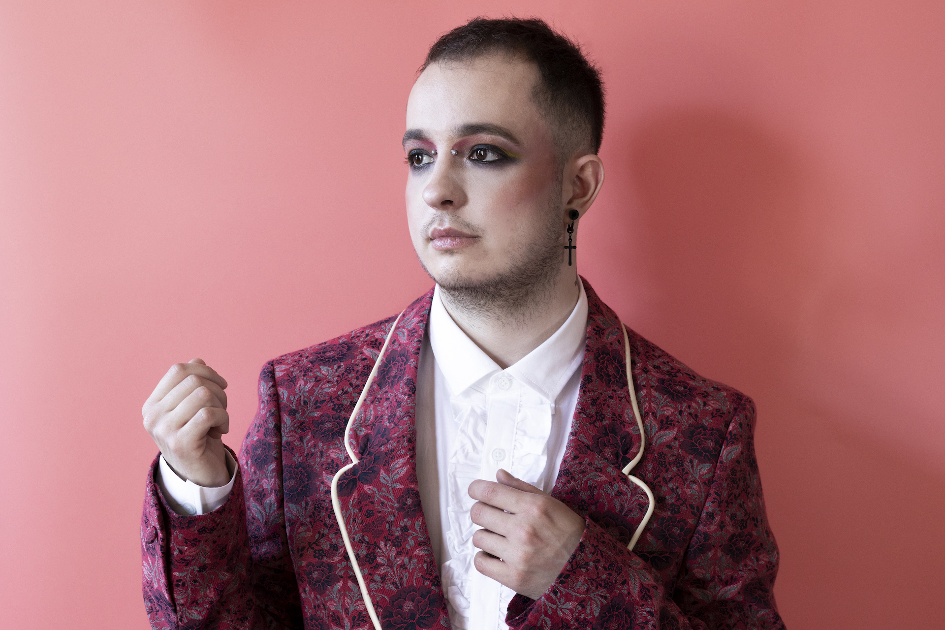 HUSK on a light pink background dressed in a dark pink flowered blazer and a white shirt looking off to the side with their hands raised around their torso.