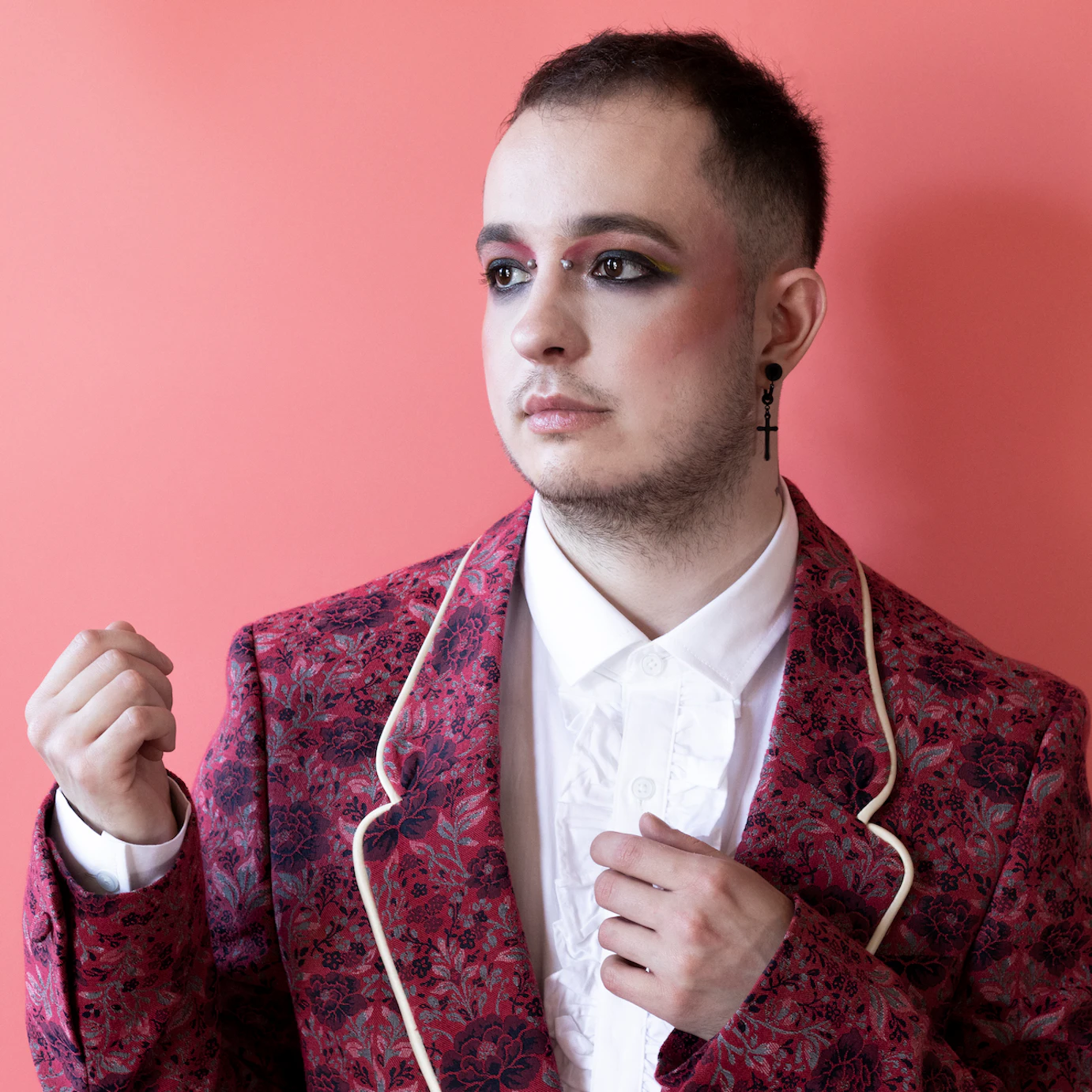 HUSK on a light pink background dressed in a dark pink flowered blazer and a white shirt looking off to the side with their hands raised around their torso.