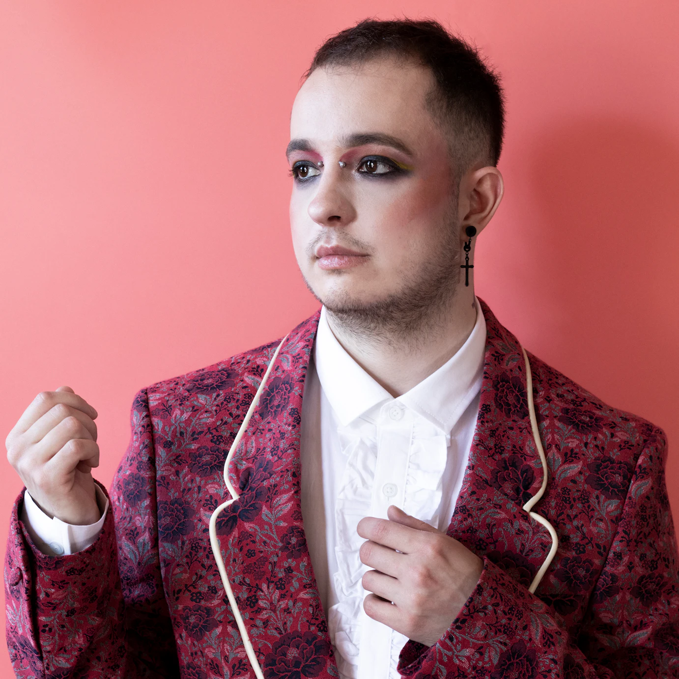 HUSK on a light pink background dressed in a dark pink flowered blazer and a white shirt looking off to the side with their hands raised around their torso.