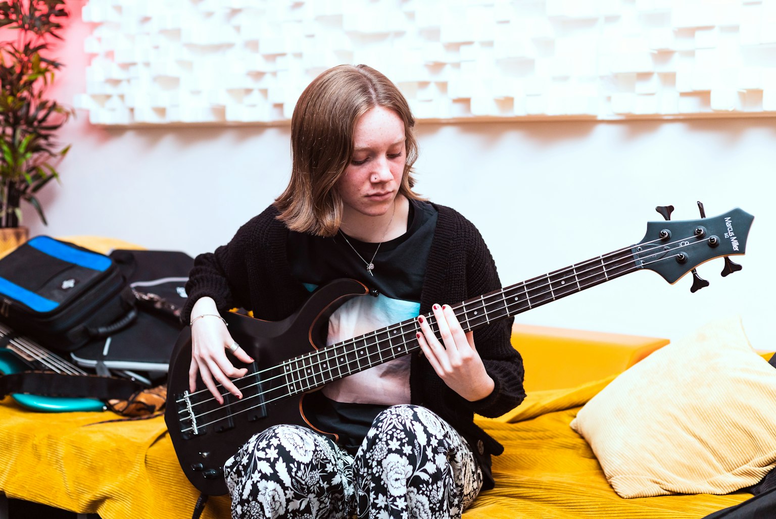 A young musician sitting on a yellow sofa playing bass guitar.