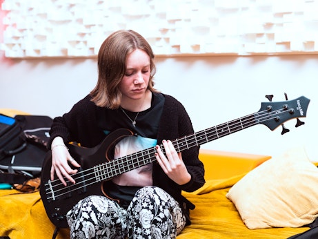 A young musician sitting on a yellow sofa playing bass guitar.