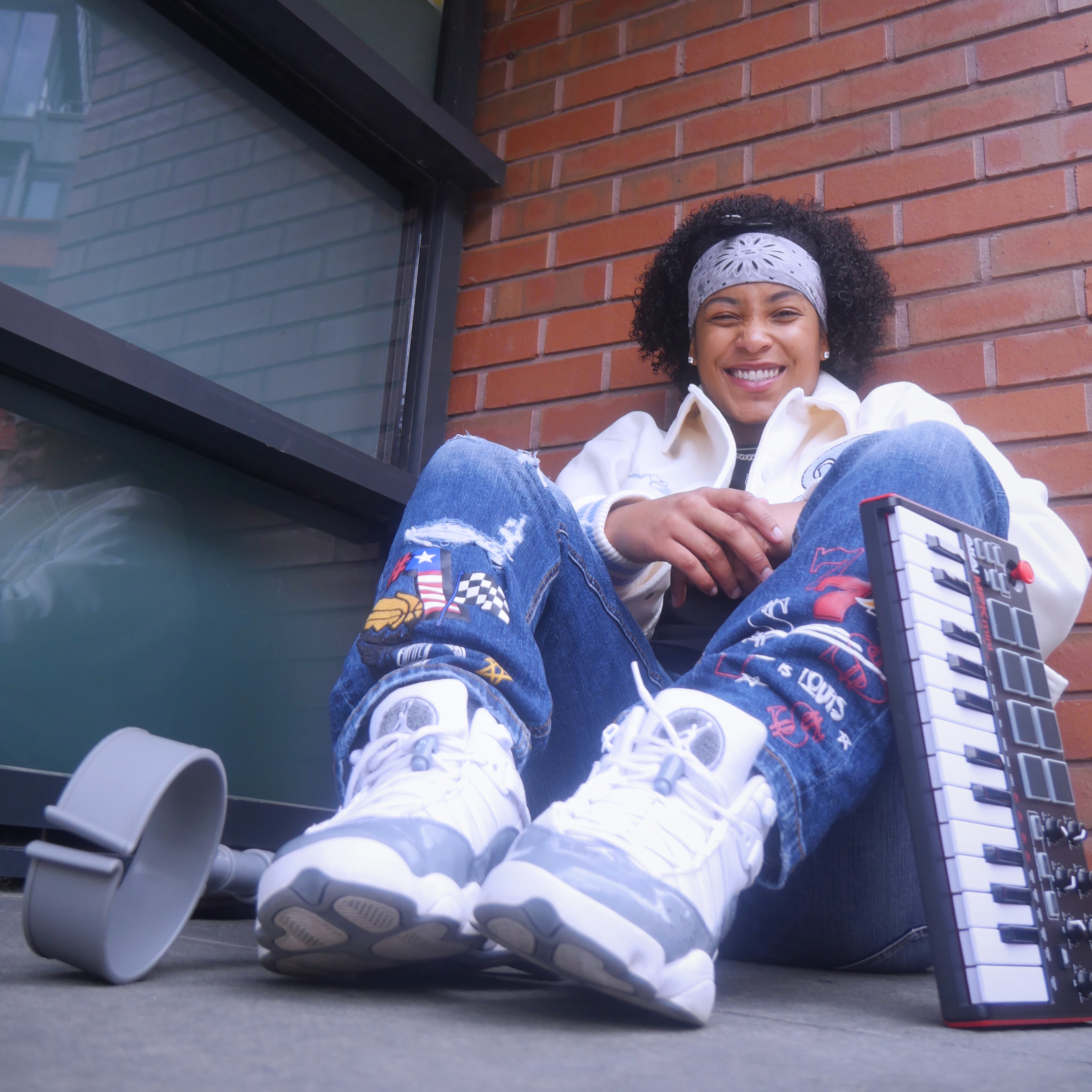 FREEQUENCY3 sat against a brick wall smiling. To her right is a crutch. To her left a MIDI keyboard.