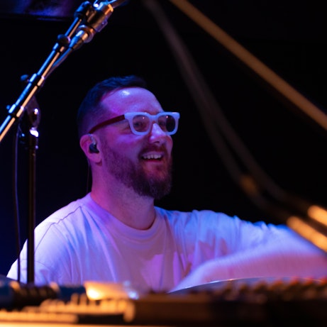 Phill Howley on stage smiling surrounded by blurred out instruments such as a keyboard and a microphone.