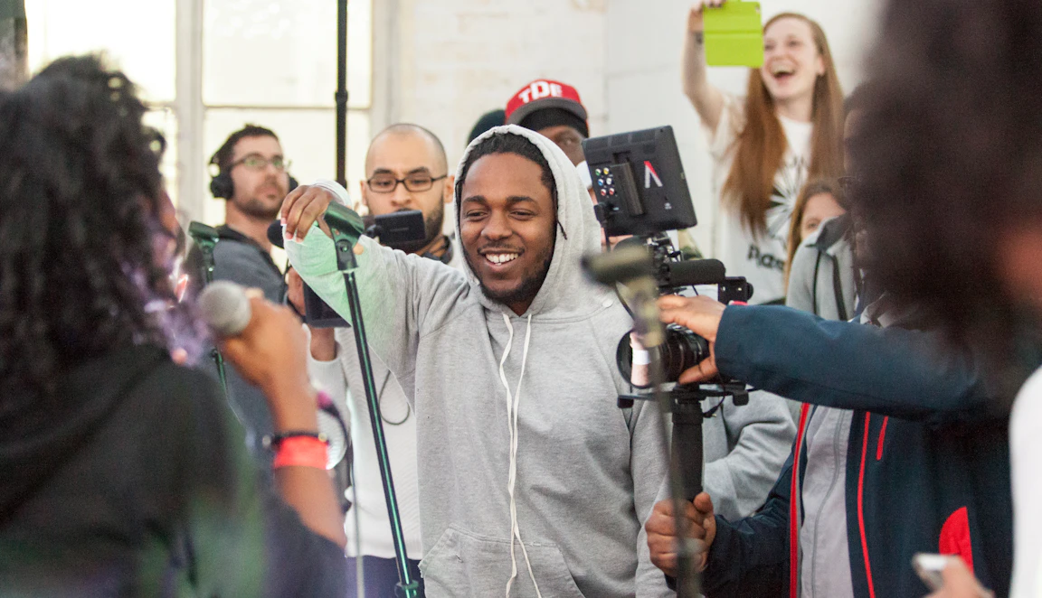 Kendrick Lamar is surrounded by a crowd of people. There is excitement in the room. A person in the background is taking a photo with their phone and has a big smile.