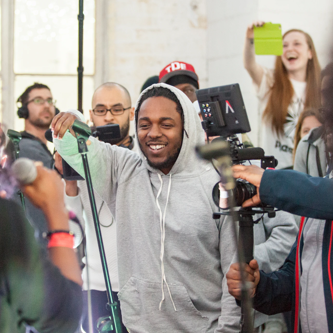 The artist Kendrick Lamar is surrounded by a crowd of people. There is excitement in the room. A person in the background is taking a photo with their phone and has a big smile.