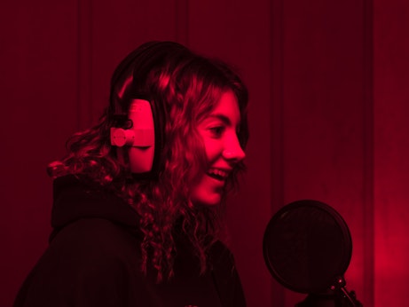 A young musician with headphones on in a recording studio smiling by a microphone.The studio is lit by deep red lighting.