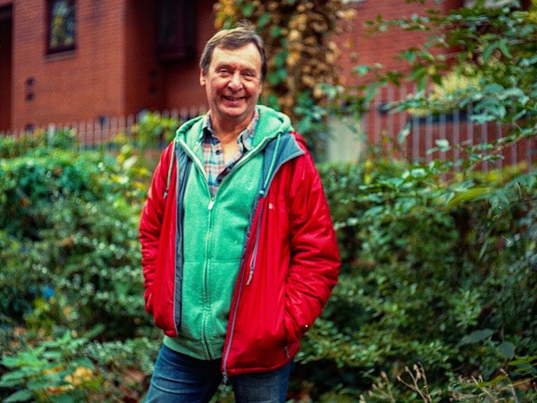 Tim is smiling, wearing a red coat and standing in front of green trees and bushes. 