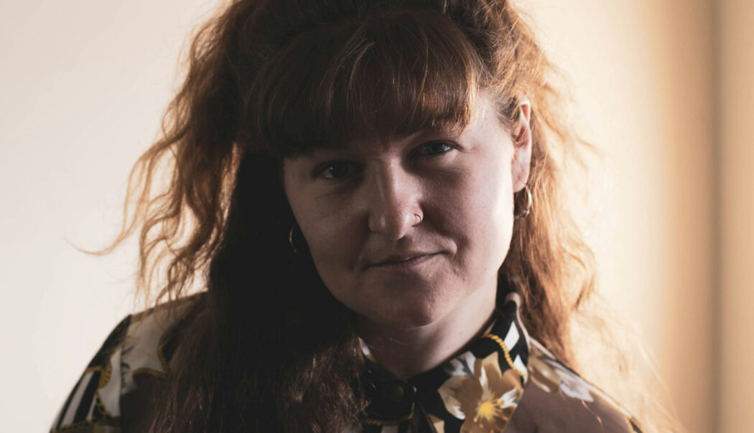 Holly Phelps (also known as IORA) stood looking into the camera on a beige background that's out of focus. She is wearing a black, white and brown button up shirt with flowers on it. Her wavy brown hair flows down the right side of her torso.