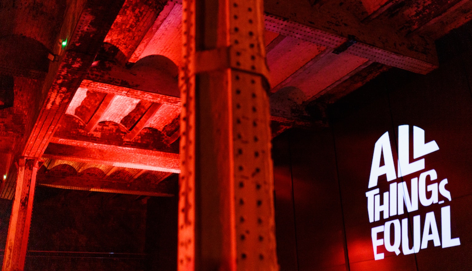 A warehouse ceiling with a metal beam in the middle of the shot, another off to the left. To the right on the wall is a projections of the All Things Equal logo. The room is lit up with a red glow.
