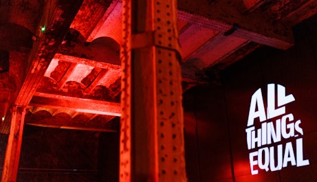 A warehouse ceiling with a metal beam in the middle of the shot, another off to the left. To the right on the wall is a projections of the All Things Equal logo. The room is lit up with a red glow.