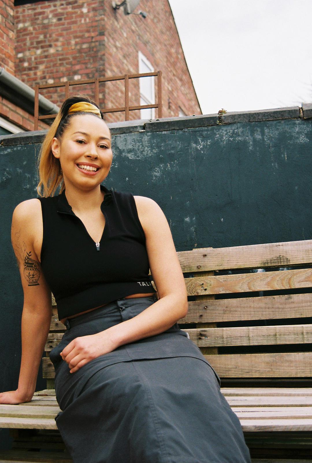 CHARMS sat on a wooden bench smiling into the camera. Behind her is the top half of a brick building.