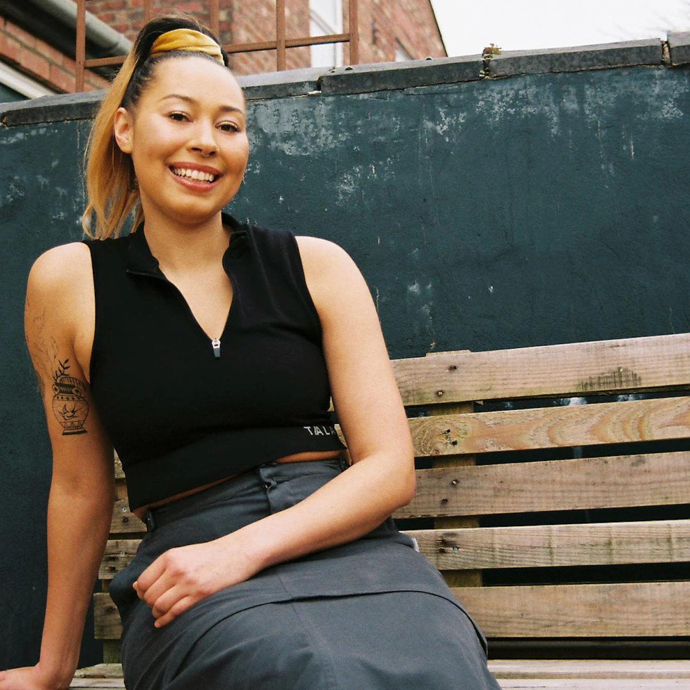 CHARMS sat on a wooden bench smiling into the camera. Behind her is the top half of a brick building.