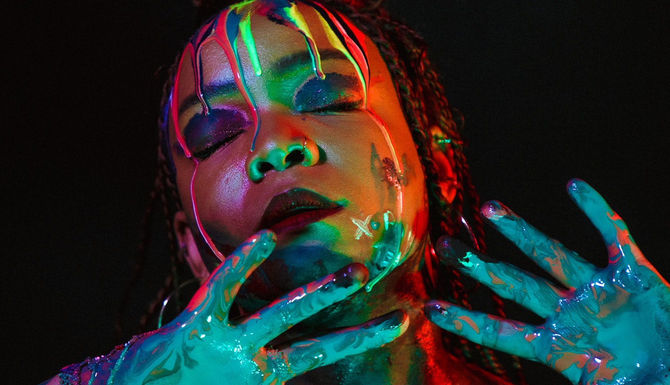 Artist Lebo, covered in multicoloured paint, on a black background. Her eyes are shut and her hands are outstretched in front of her.