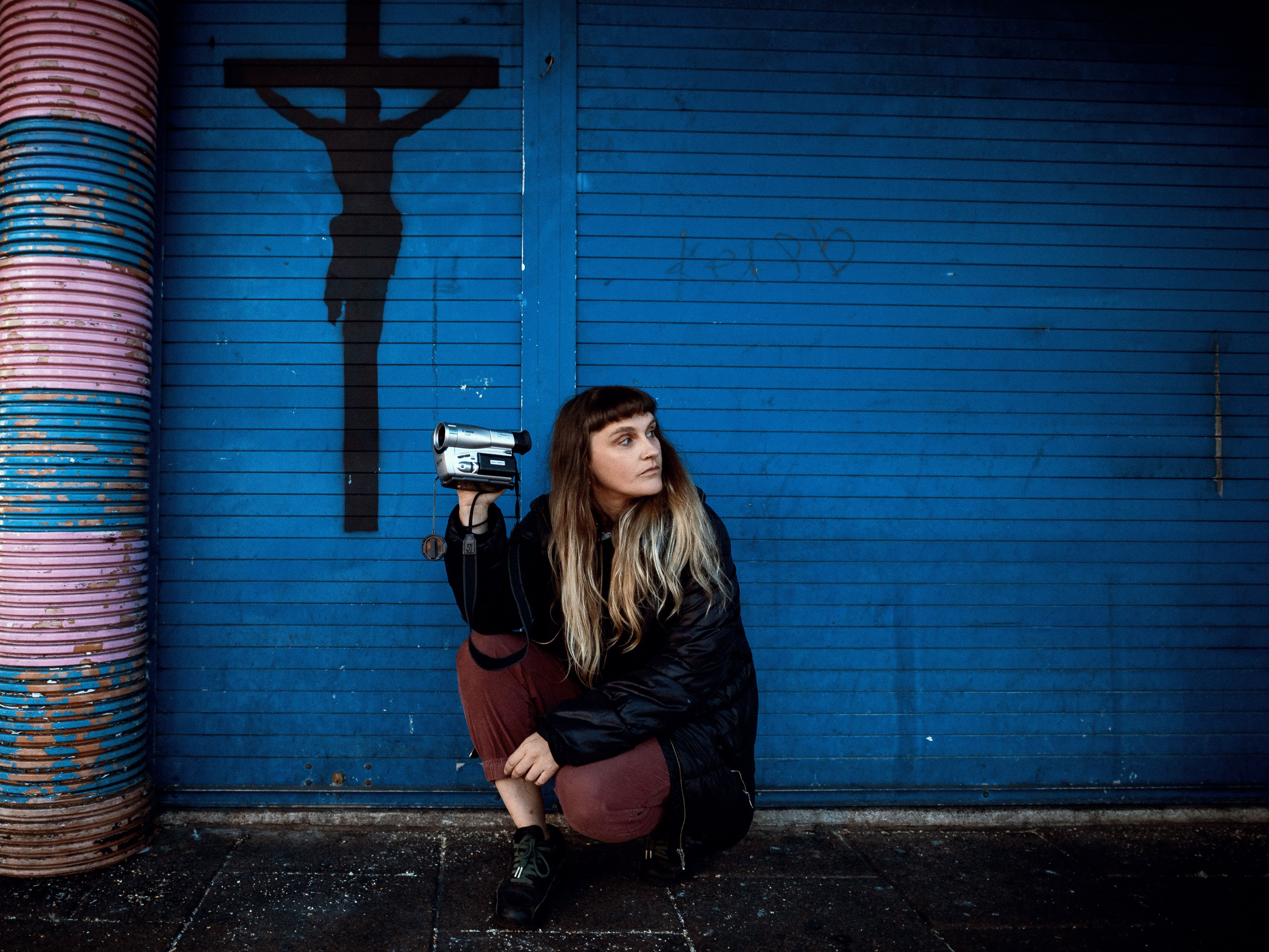 MARF is crouched beside blue shutters with a crucifix spray painted in black on the left hand side. She's holding a video recorder in one direction and facing the other.