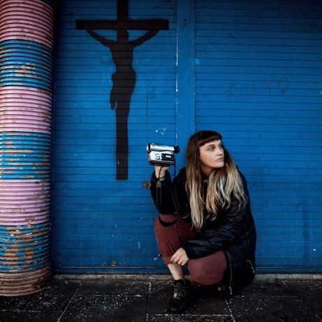 MARF is crouched beside blue shutters with a crucifix spray painted in black on the left hand side. She's holding a video recorder in one direction and facing the other.