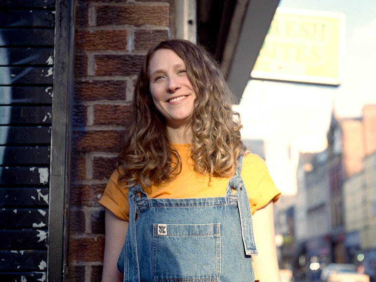 Lucy stands against a brick wall on a sunny street in Manchester. She is smiling and look directly at the camera. She has mid-length curly brown hair and is wearing denim dungarees.