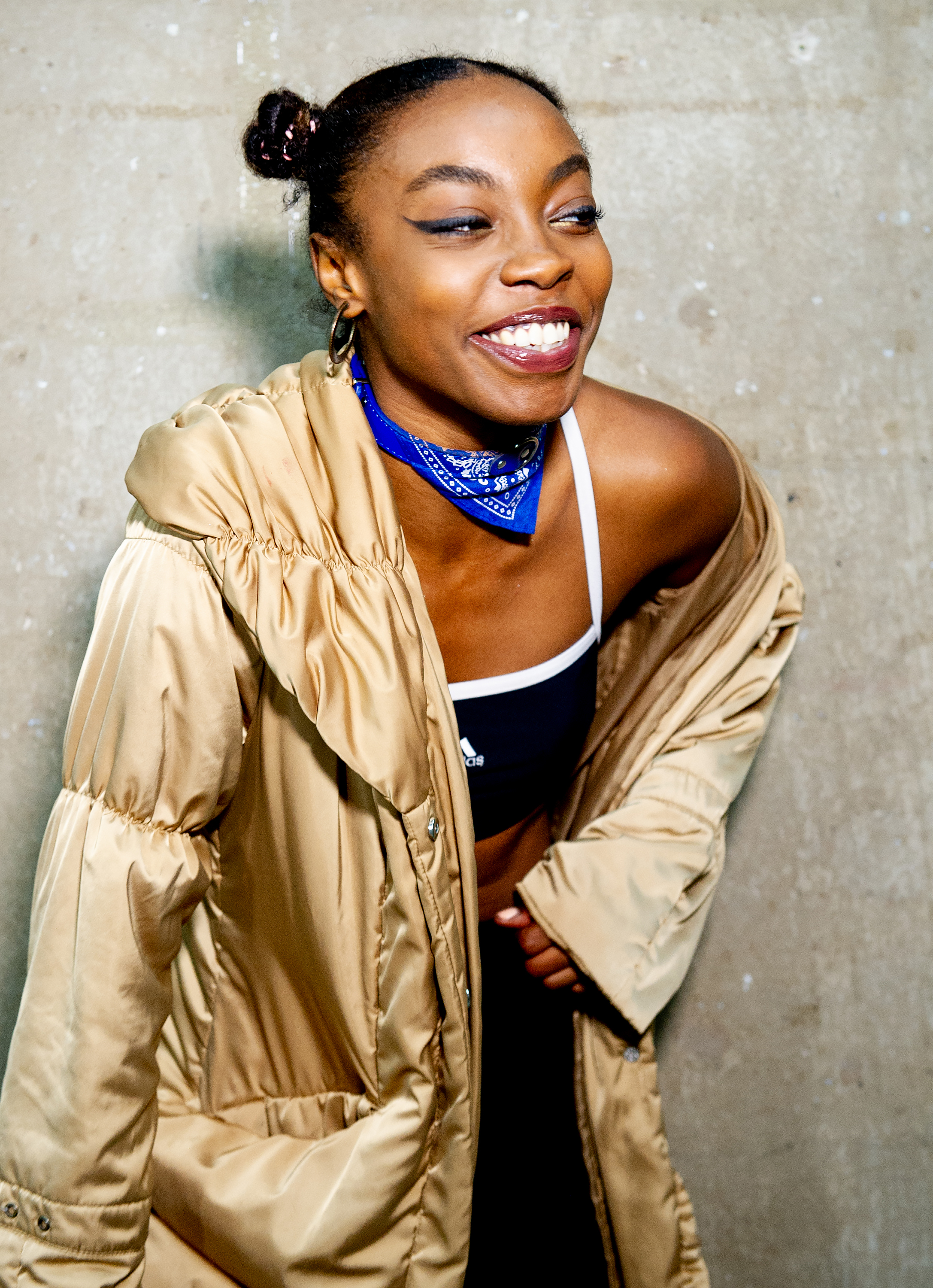 Sarah Angel smiling and looking off to the right. She's wearing a long golden coat and a blue and white bandana wrapped around her neck.