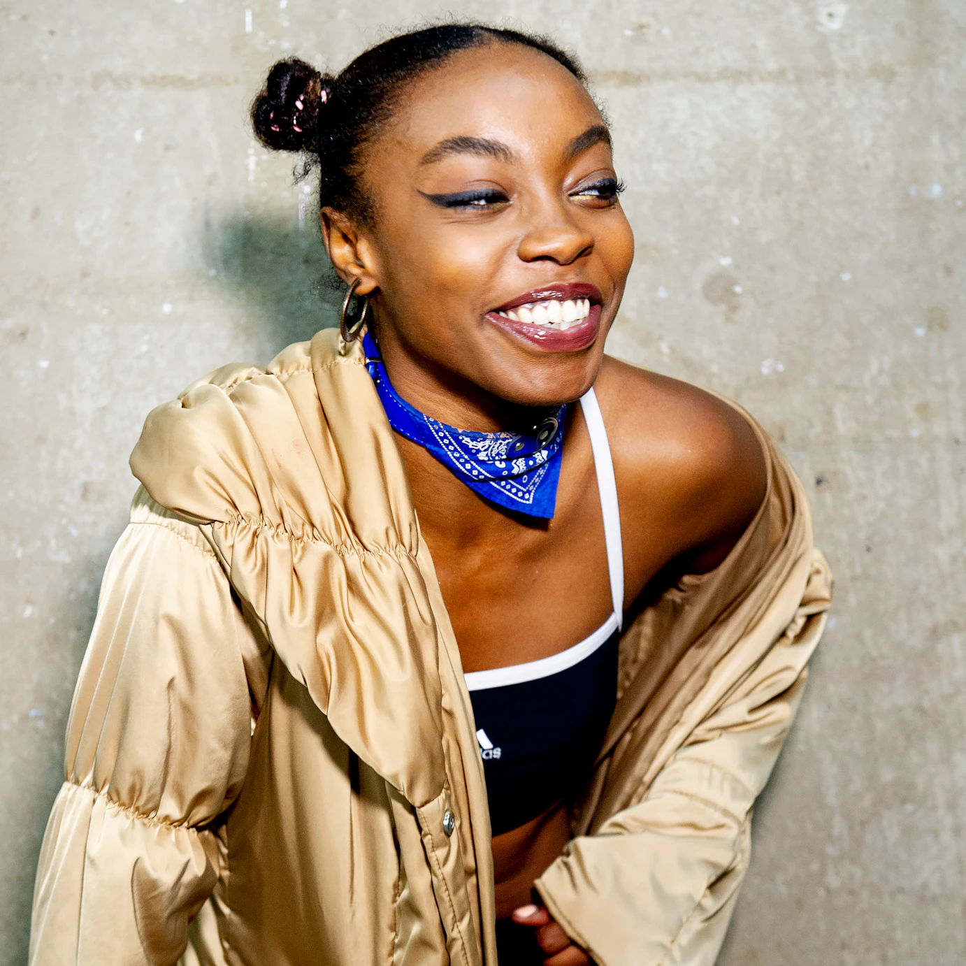 Sarah Angel smiling and looking off to the right. She's wearing a long golden coat and a blue and white bandana wrapped around her neck.