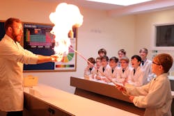 Students wearing protective gear in Science class with their teacher. The teacher is conducting an experiment with a student which is producing a small ball of fire, whilst the other students watch excitedly.