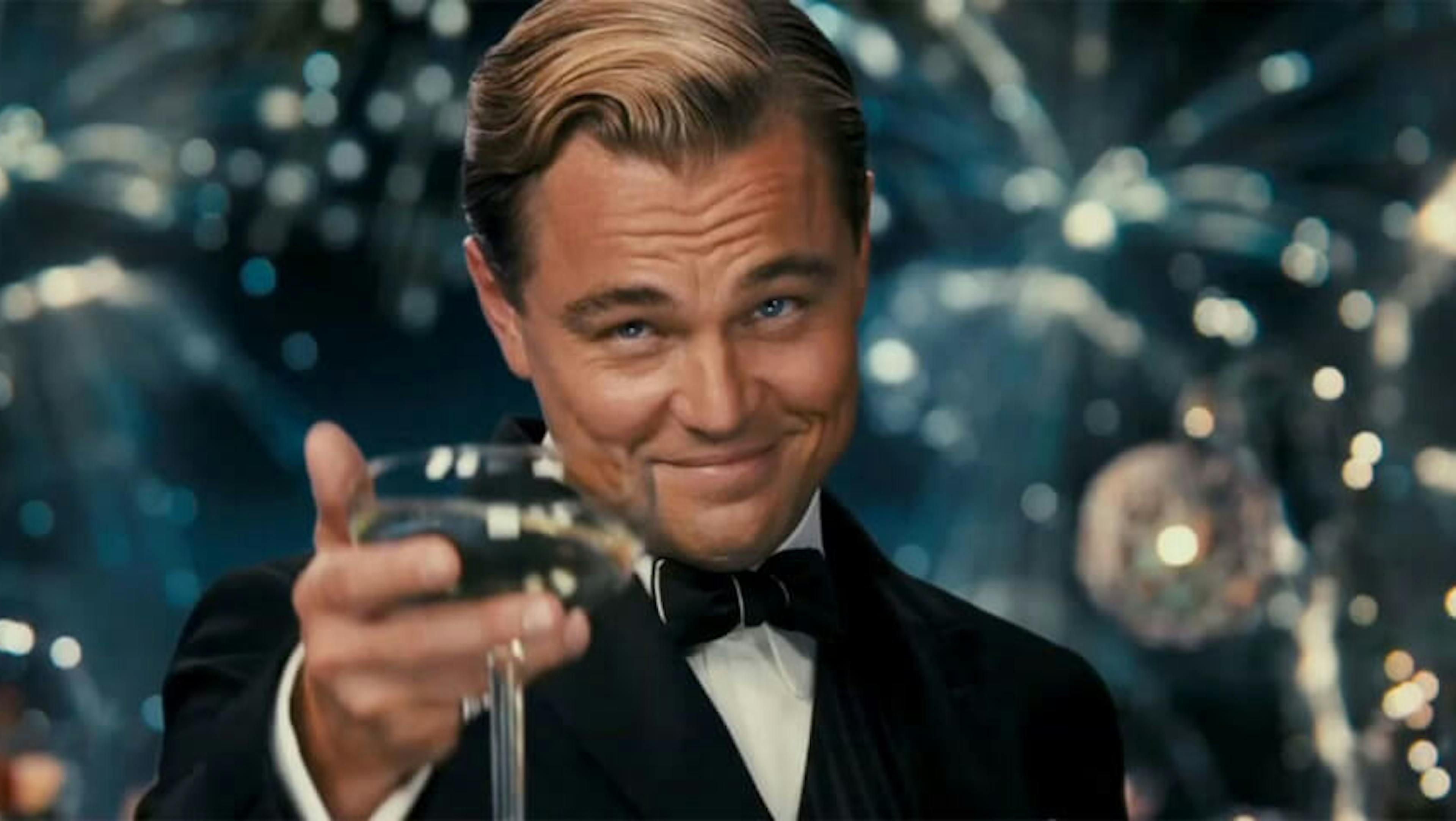 Cena do filme Gatsby onde ele oferece uma taça de champagne sorrindo e com fogos de artifício ao fundo