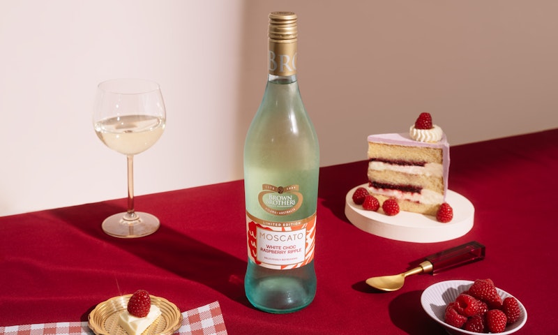 A bottle of Moscato White Choc Raspberry Ripple styled with a jam & cream layer cake, wine, and bowl of raspberries against a deep red and white backdrop