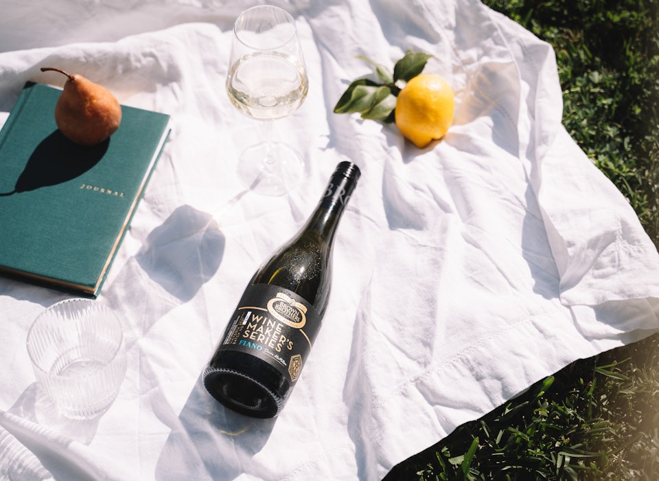 Brown Brothers Winemaker's Series Fiano bottle laying on a picnic rug with a book, glass and fruit
