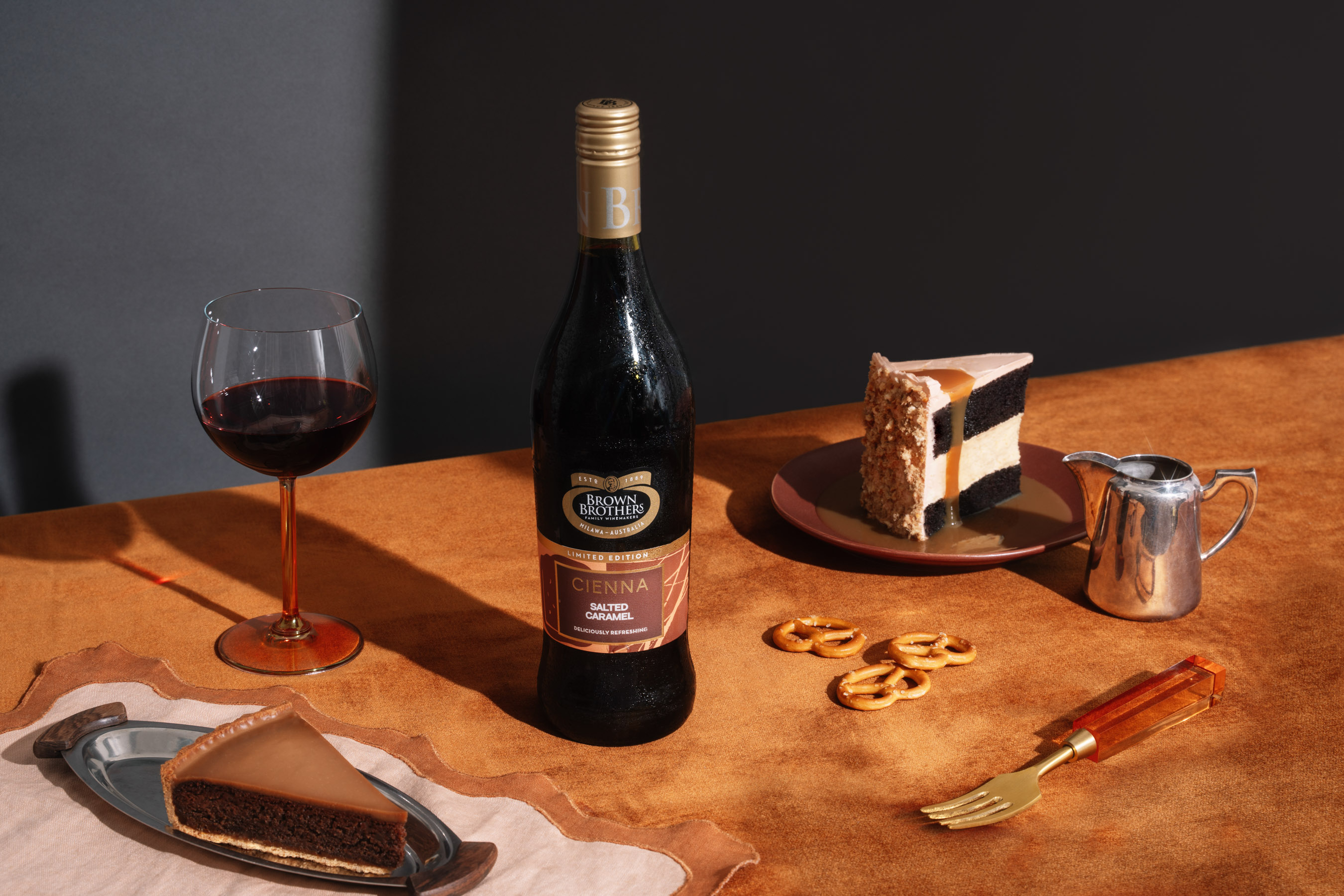 A bottle of Cienna Salted Caramel styled with a decadent slice of cake, caramel drizzle and salted pretzels against a richly coloured backdrop of moody blue and burnt orange 