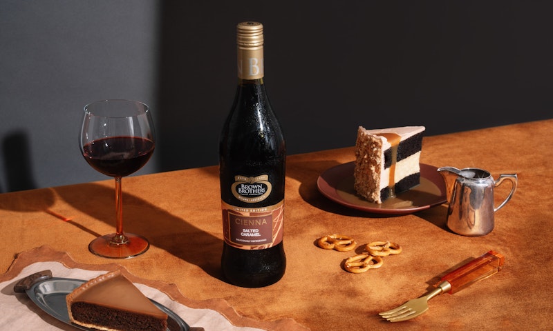 A bottle of Cienna Salted Caramel styled with a decadent slice of cake, caramel drizzle and salted pretzels against a richly coloured backdrop of moody blue and burnt orange 