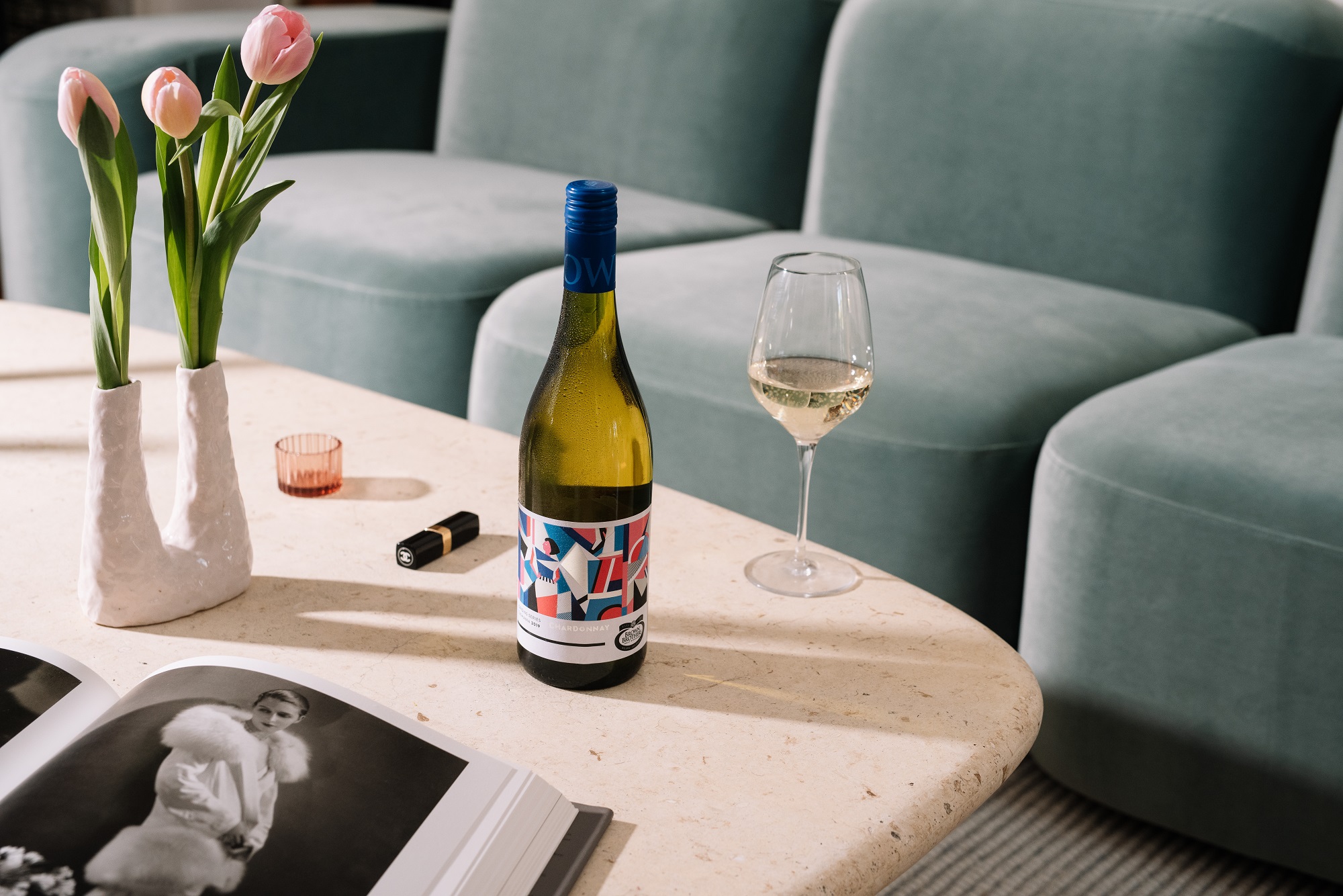 bottle of Origins Series Chardonnay on a stone coffee table set with pink tulips, lipstick, a fashion book and wine glass