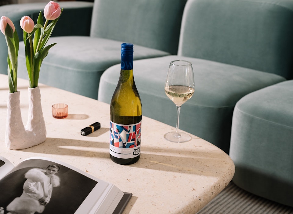 bottle of Origins Series Chardonnay on a stone coffee table set with pink tulips, lipstick, a fashion book and wine glass