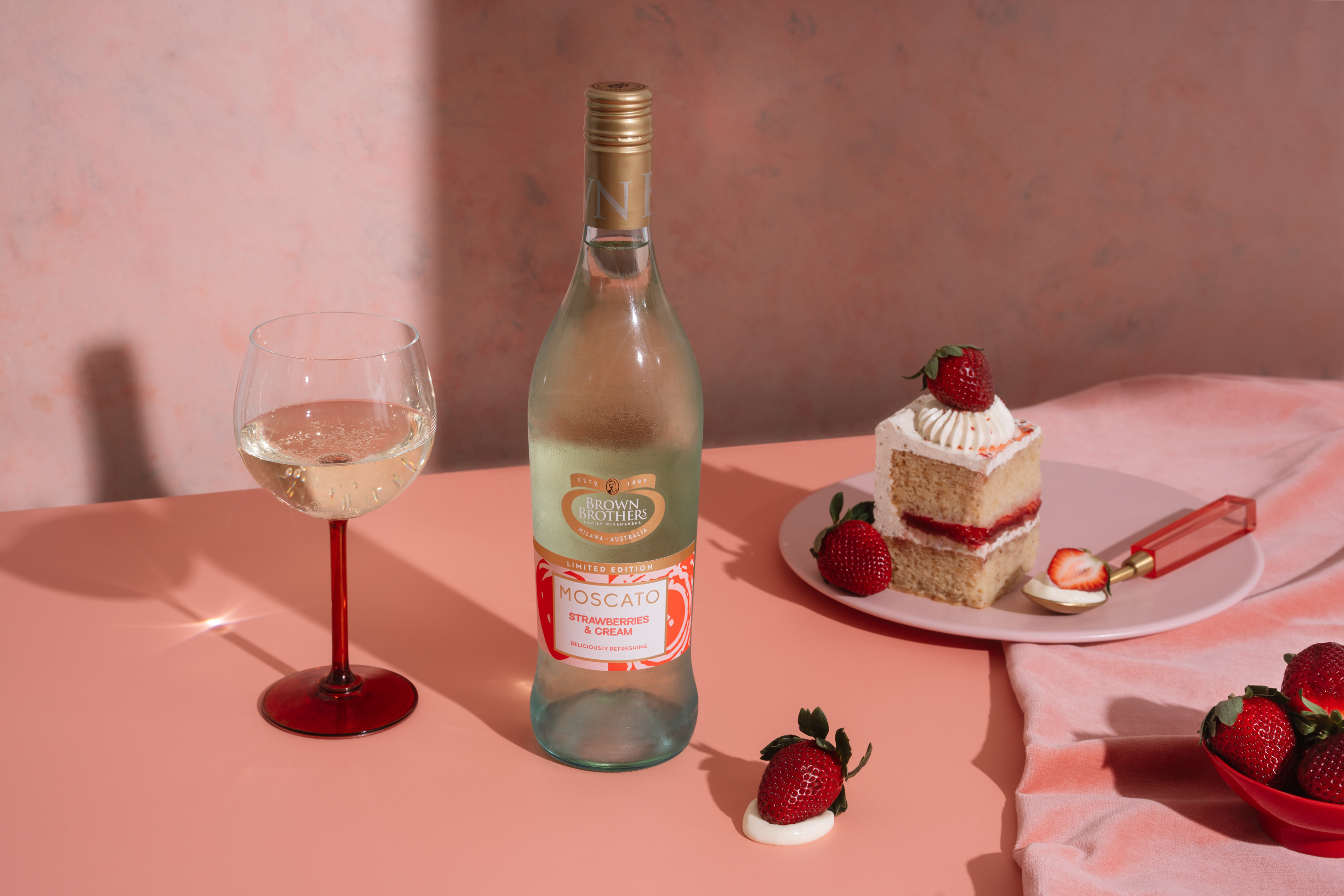 A bottle of Moscato Strawberries & Cream styled with cake, strawberries, and wine against a beautiful pink backdrop.