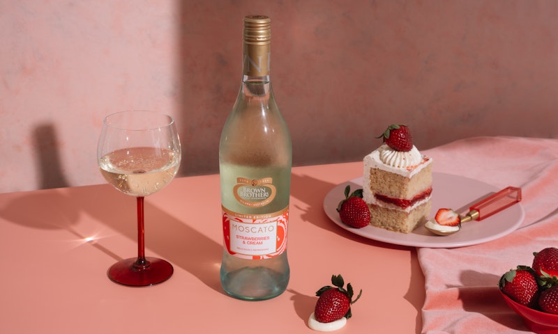 A bottle of Moscato Strawberries & Cream styled with cake, strawberries, and wine against a beautiful pink backdrop.