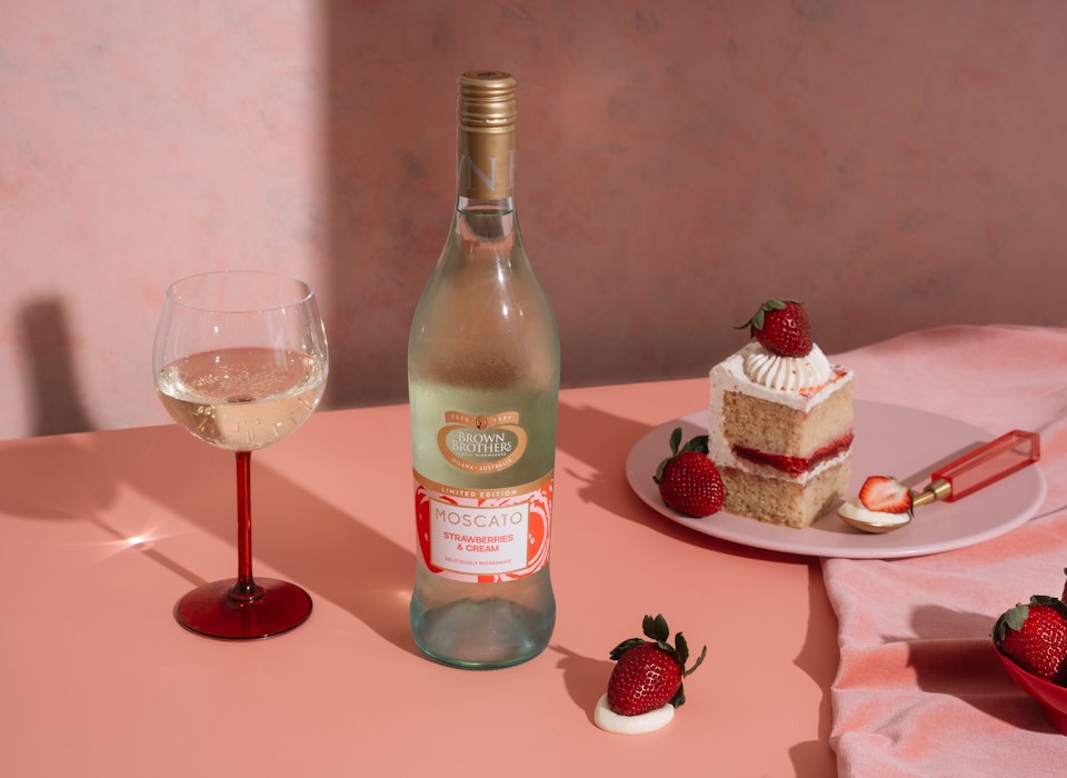 A bottle of Moscato Strawberries & Cream styled with cake, strawberries, and wine against a beautiful pink backdrop.