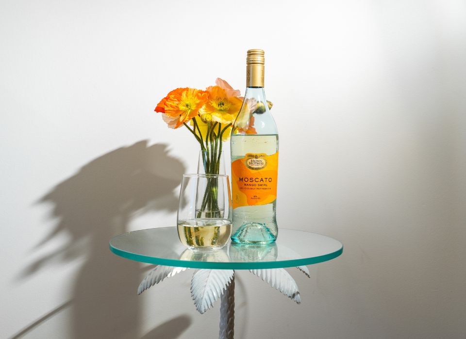 A bottle of Brown Brothers Moscato Mango Swirl on a table with a flower vase