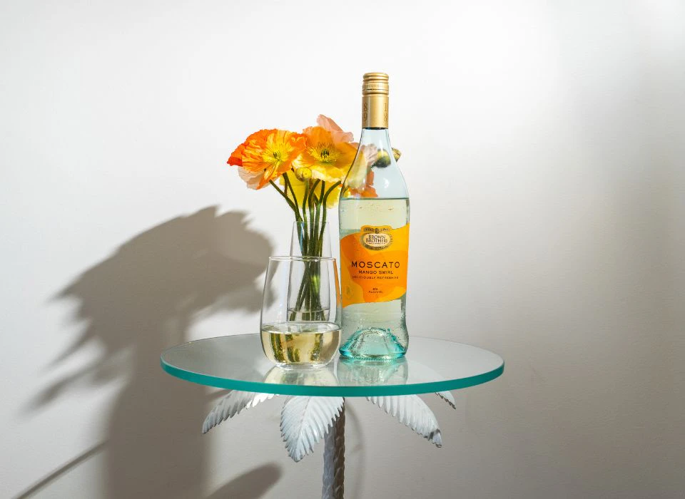 A bottle of Brown Brothers Moscato Mango Swirl on a table with a flower vase