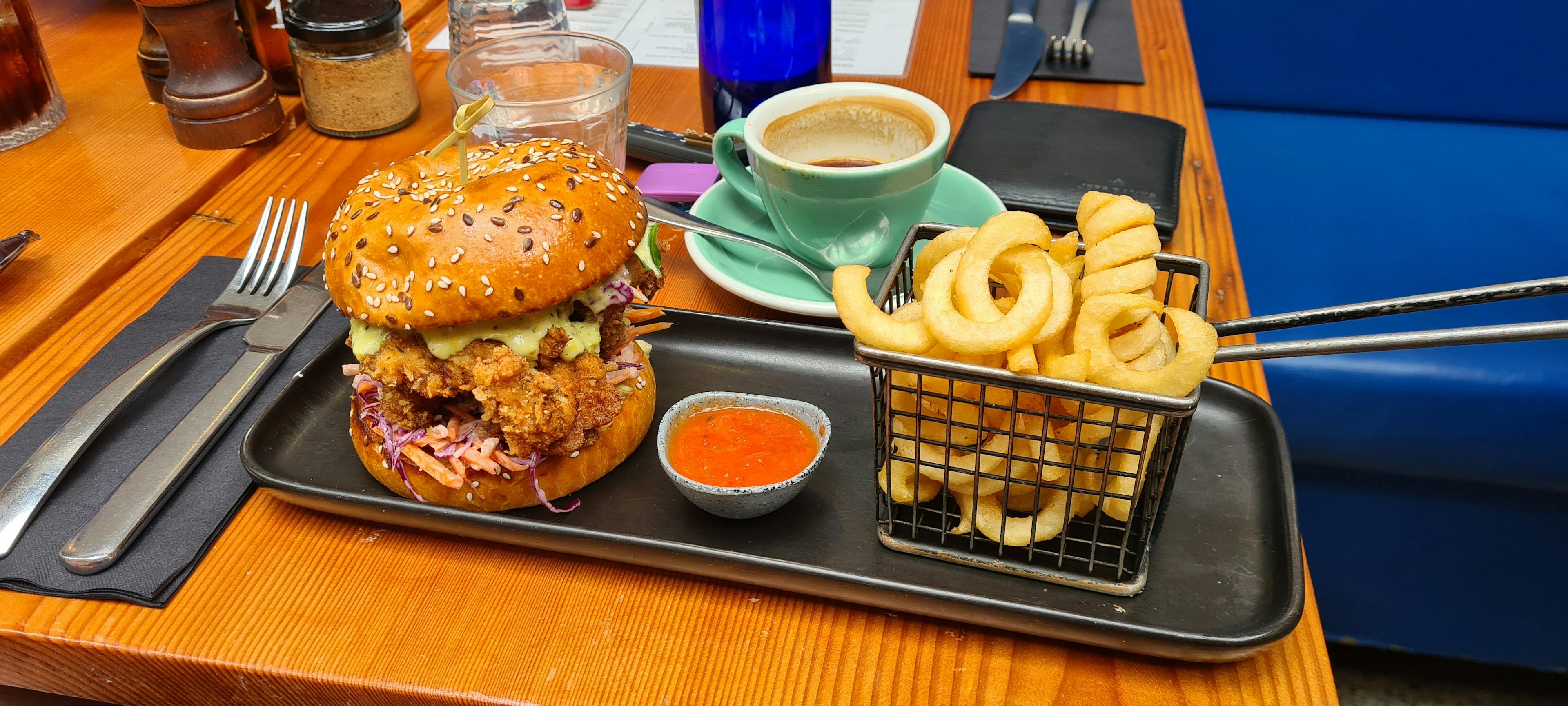 Seeded burger bun with crispy fried chicken, slaw and sauce, next to a basket of curly fries.