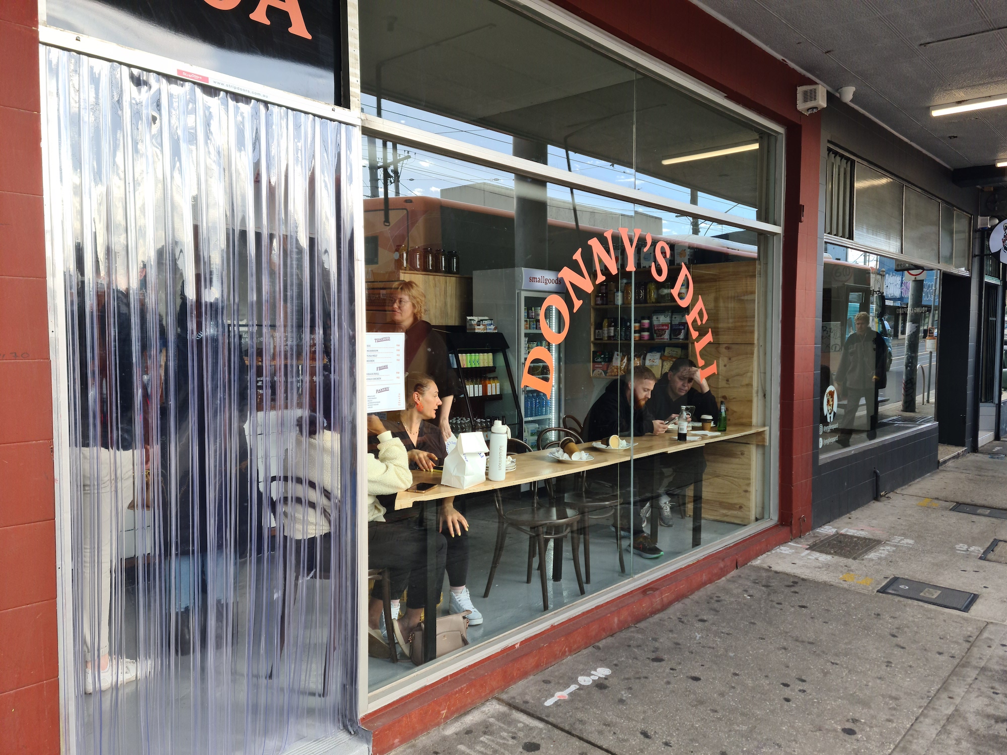 People sitting at bench facing a large floor to ceiling window at the front Donny's Deli. The window has a decal with the business' name on it.