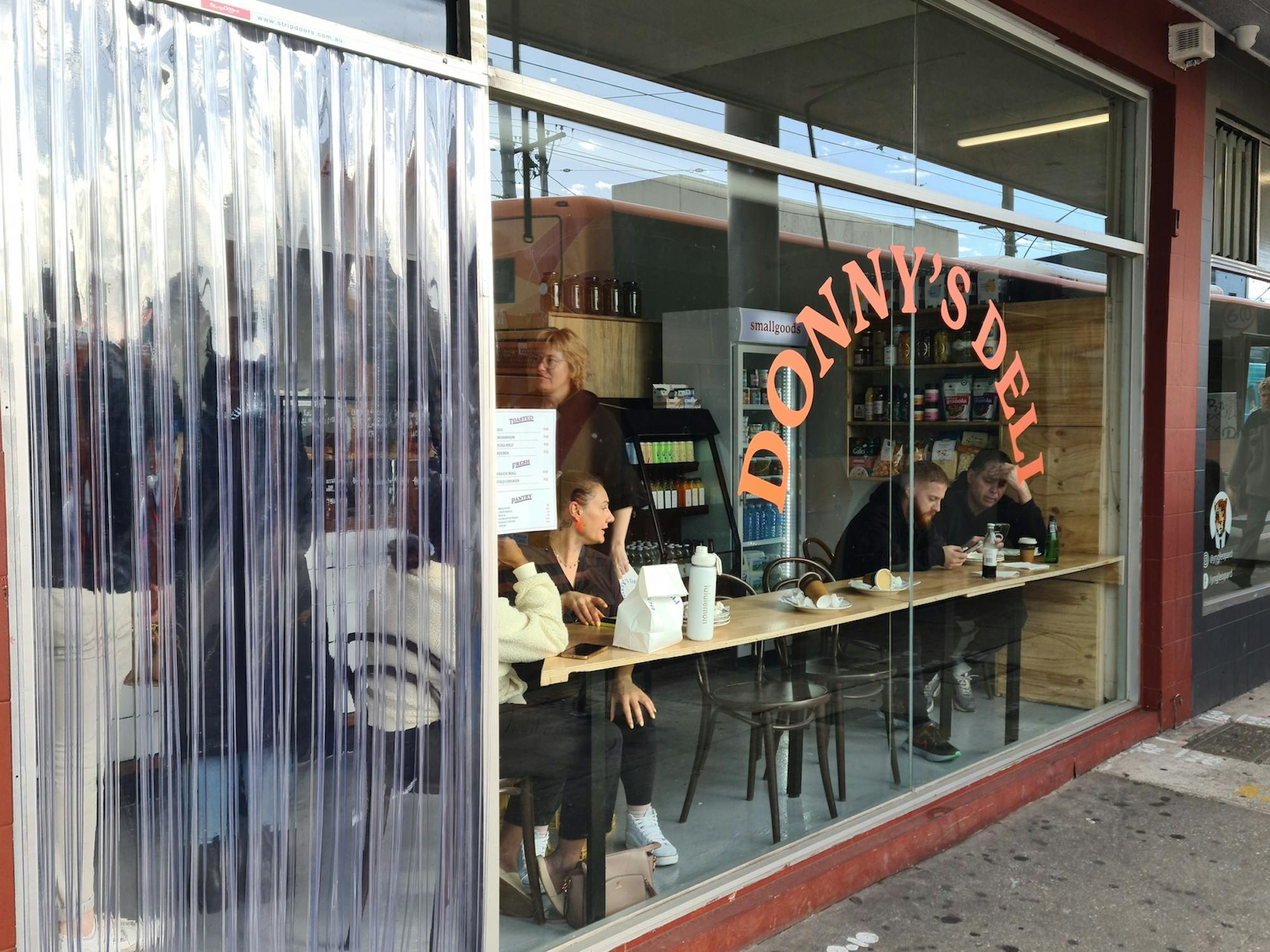 People sitting at bench facing a large floor to ceiling window at the front Donny's Deli. The window has a decal with the business' name on it.